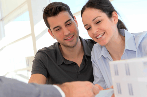 Una pareja sonriente está sentada ante un escritorio, revisando documentos con una persona vestida de traje, lo que sugiere una reunión de negocios o financiera, posiblemente sobre bienes inmuebles o un contrato.